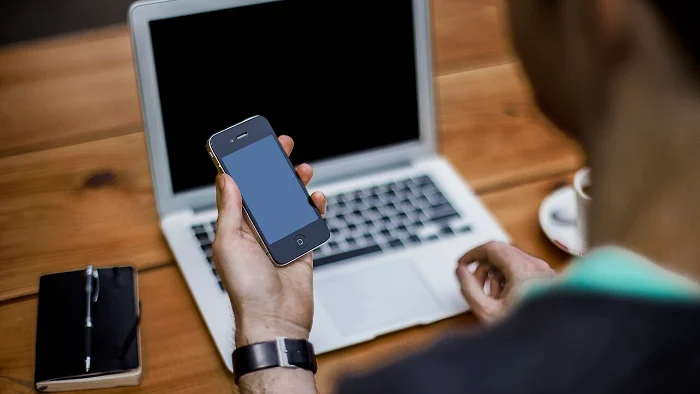 Person mit Smartphone in der Hand vor einem aufgeklappten Laptop